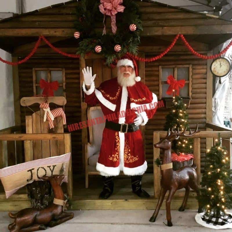 Santa waving in a log-cabin doorway with wooden reindeer figures.