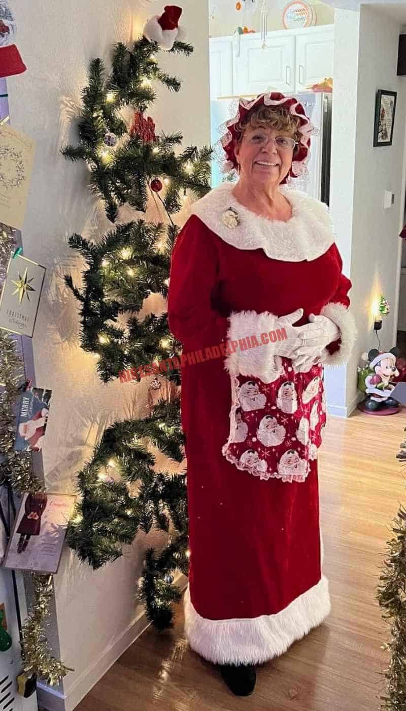 Mrs. Claus for hire in Philadelphia standing beside a slim lit tree in a home hallway.