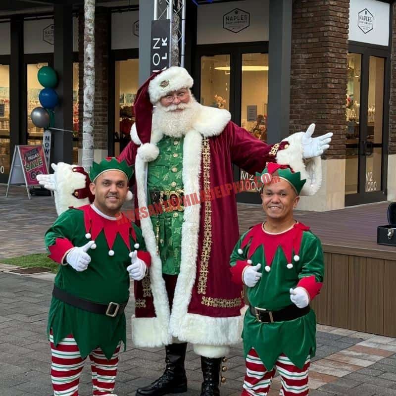 Santa standing with two elves in green costumes, thumbs-up.